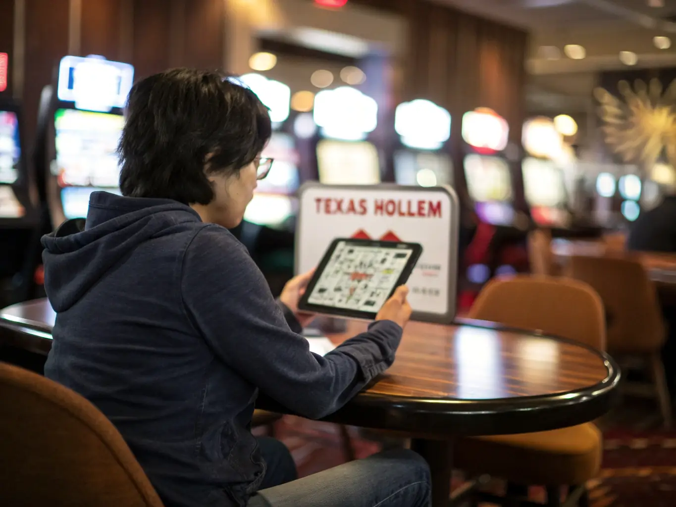 A person is interacting with a live dealer on the GBET app on their smartphone while at a coffee shop. The app interface shows a live video stream of the dealer and the table game.